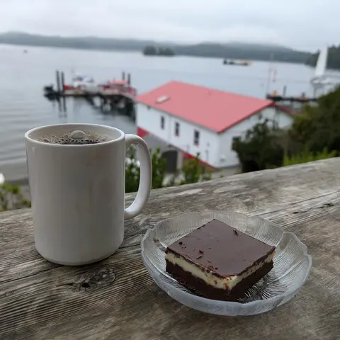 A white ceramic mug of black coffee next to a nanaimo bar on a glass plate, resting on a weathered wooden railing. Behind them, a red-roofed white boathouse sits on the water in a misty harbor, with sailboats and forested hills in the distance.
