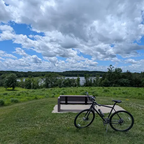 A dark grey bicycle on its kickstand in mown grass in front of an empty wooden bench on a concrete pad, with a meadow, a line of trees, a river, and tall clouds behind them.