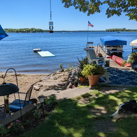 A lake seen from a cabin yard on a bright day, with a sand beach and kids' toys in the foreground, a turquoise floating mat and an inner tube in the shallow water, a dock with a pontoon boat, patio chairs under a blue umbrella, and a black and white greyhound standing on the grass at the edge of the frame.