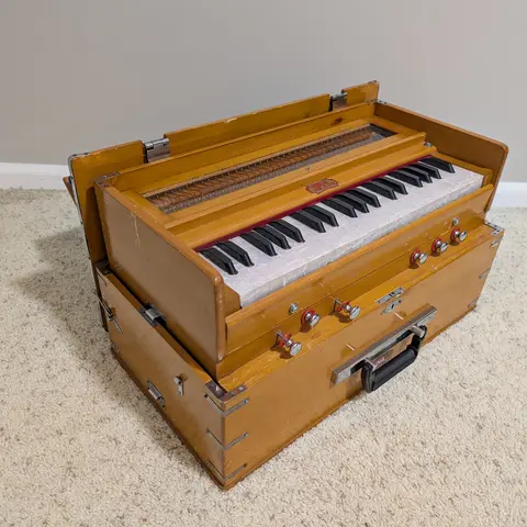 A wooden harmonium sitting open on beige carpet in the corner of a room, its top panel raised above the black and white keyboard, with a row of chrome and red stop knobs and a carrying handle along the front.