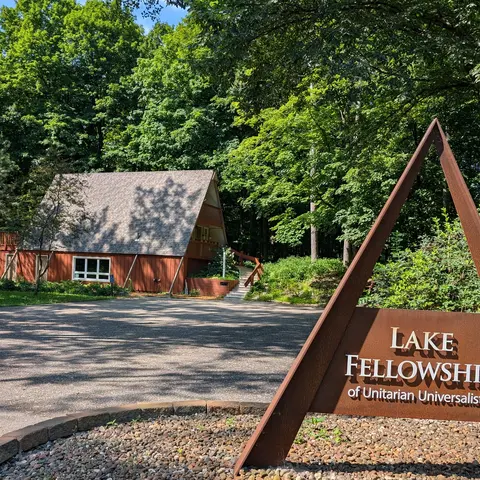 A triangular rusted-metal sign in the foreground reading "Lake Fellowship of Unitarian Universalists" with a wooden A-frame building nestled in lush green woods in the background.