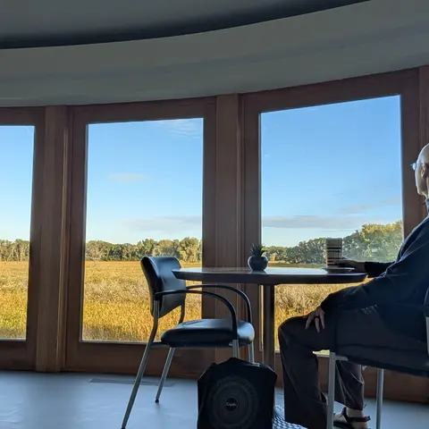 A person in a hoodie sits at a small round table by a curved bank of windows, mug in hand, looking out over a golden marsh toward a tree line under a clear sky.