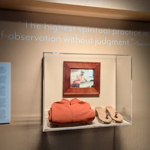 A quote painted on a wall reading "The highest spiritual practice is self-observation without judgment" above a glass display case holding a framed photo of Swami Kripalu at a writing desk, a folded orange robe, and a pair of wooden sandals, with a text panel to the left.