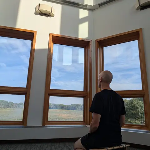 A bald man named Matthew in a black T-shirt and shorts kneels barefoot on a low wooden meditation bench, seen from behind, facing tall wood-framed windows that look out over a marsh and treeline under a blue sky, with sunlight falling across the white wall above.