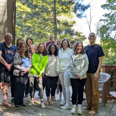 Thirteen people stand together on a wooden deck outside a small A-frame building, with trees behind them.