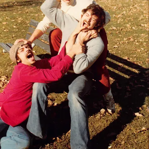 Three men beside a park bench, grabbing one another, sticking out their tongues, and making exaggerated faces.
