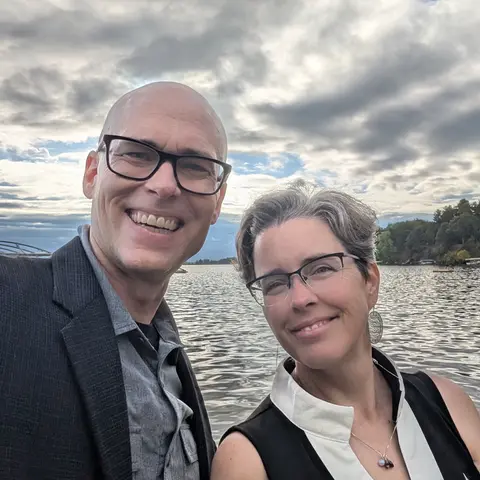 Matthew and Marnie smiling in a selfie on the shore of a lake, with a cloudy sky behind them and trees along the far bank