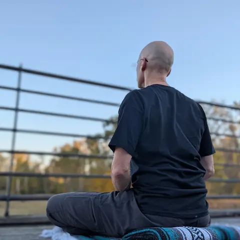 Matthew sitting cross-legged on a folded blanket on The Marsh deck, seen from behind, with autumn trees and blue sky beyond the railing.