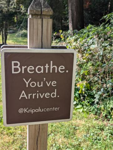 A sign with a brown backgroud and tan letters that reads Breathe. You've Arrived.