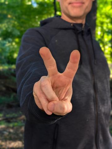 Matthew standing in the woods with blue sweatshirt holding out a peace sign