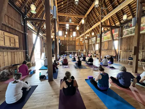 About thirty people sitting cross-legged on mats on a wood floor inside a timber-framed barn, facing a teacher seated at the far end, daylight coming through the open door.
