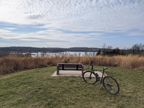 a bench on the top of a hill overlooking grass and a lake in a park on a cloudy day