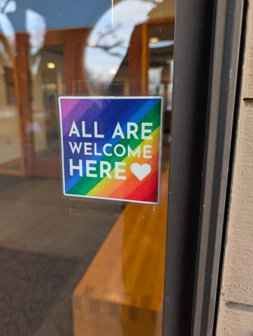 A rainbow colored sign with the words all are welcome here on a window at The Marsh