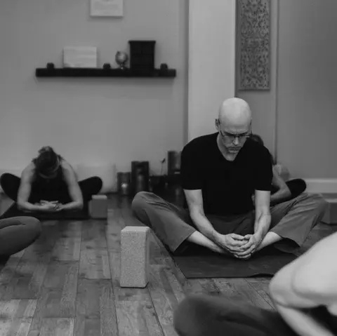 Black and white photo of Matthew on a mat with the soles of his feet together, hands clasped around his feet and head lowered, two other students on mats behind him.