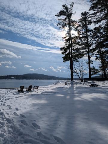 footprints in the snow toward chairs by a lake framed by tall pine trees