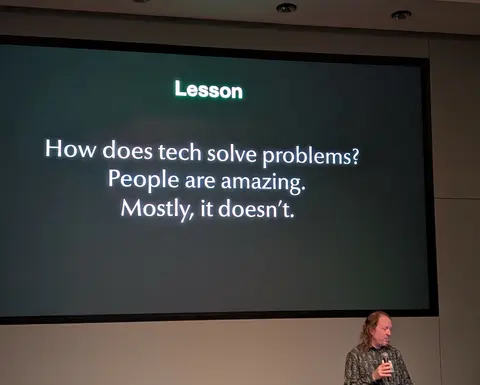 Presentation slide titled “Lesson” reading: “How does tech solve problems? People are amazing. Mostly, it doesn’t.” with a speaker holding a microphone in the lower right.