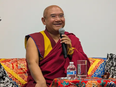 A smiling, shaved-headed Tibetan Buddhist monk wearing traditional maroon and yellow robes. He is wearing glasses and holding a black microphone in his right hand, around which a strand of yellow prayer beads (mala) is wrapped. He is seated on a couch with colorful, ornate, patterned fabric. In front of him, on a small table draped in a matching patterned cloth, sits a clear plastic water bottle and a glass of water. The background is a plain white wall.
