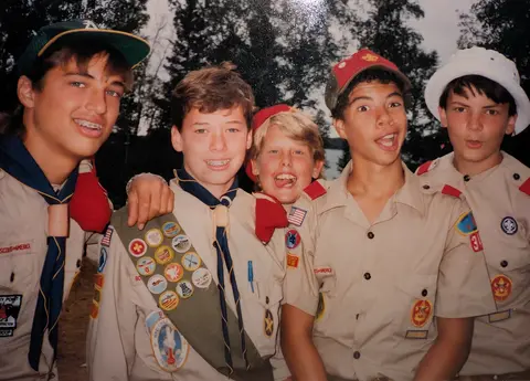 A grainy vintage photograph of five young Boy Scouts in tan uniforms posing closely together outdoors. The second boy from the left wears a merit badge sash and smiles at the camera, while a blonde boy peeks over his shoulder making a goofy face with his tongue out. The other boys wear various hats and neckerchiefs, posing with expressive, wide-eyed faces against a background of trees and water.