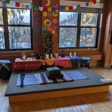 A yoga mat and blanket laid out on a wooden platform in front of an altar with candles, a framed photo, carved figures, and hand-decorated prayer flags, in a sunlit room with snow visible outside the windows.