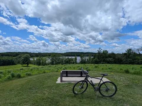 A dark grey bicycle on its kickstand in mown grass in front of an empty wooden bench on a concrete pad, with a meadow, a line of trees, a river, and tall clouds behind them.