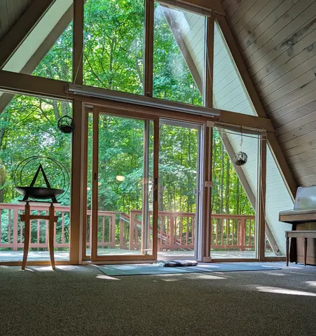 Interior of an A-frame room with large glass windows and sliding doors opening to a wooden deck surrounded by lush green trees, featuring a yoga mat on the carpeted floor, a upright piano on the right, and a decorative bowl stand on the left.