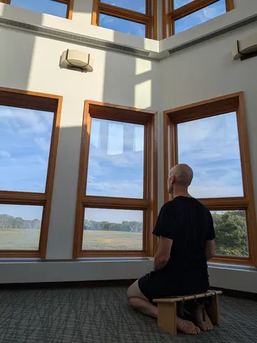 A bald man named Matthew in a black T-shirt and shorts kneels barefoot on a low wooden meditation bench, seen from behind, facing tall wood-framed windows that look out over a marsh and treeline under a blue sky, with sunlight falling across the white wall above.