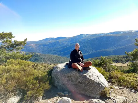Matthew smiling and sitting cross-legged on a large rock with a sweeping view of a mountainous Spanish landscape and green valley.
