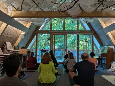 A group of people sit on yoga mats on the floor of an A-frame room, facing a bald man with glasses seated in front of tall windows, trees visible through the glass and gauzy fabric and bare branches hung across the ceiling.