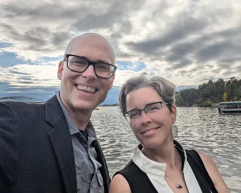 Matthew and Marnie smiling in a selfie on the shore of a lake, with a cloudy sky behind them and trees along the far bank