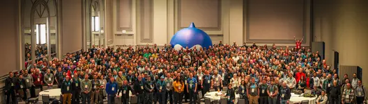 A wide-angle shot of a crowd of participants gathered in a Chicago conference hall for DrupalCon 2026, with a large, blue inflatable Druplicon mascot centered in the room.