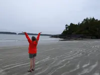 A person in a red jacket stands on a wet sandy beach with arms raised toward an overcast sky, facing the ocean