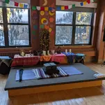 A yoga mat and blanket laid out on a wooden platform in front of an altar with candles, a framed photo, carved figures, and hand-decorated prayer flags, in a sunlit room with snow visible outside the windows.
