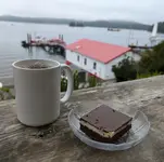 A white ceramic mug of black coffee next to a nanaimo bar on a glass plate, resting on a weathered wooden railing. Behind them, a red-roofed white boathouse sits on the water in a misty harbor, with sailboats and forested hills in the distance.
