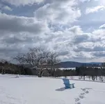 A snow-covered field at Kripalu on a bright winter day, with a single empty blue Adirondack chair, a set of footprints leading toward it, a bare tree, and clouds rolling over distant hills.