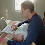A man types on a white laptop at a kitchen table, one hand on the trackpad, while a newborn in a pale blue sleeper sleeps across his forearm.
