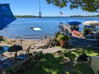 A lake seen from a cabin yard on a bright day, with a sand beach and kids' toys in the foreground, a turquoise floating mat and an inner tube in the shallow water, a dock with a pontoon boat, patio chairs under a blue umbrella, and a black and white greyhound standing on the grass at the edge of the frame.