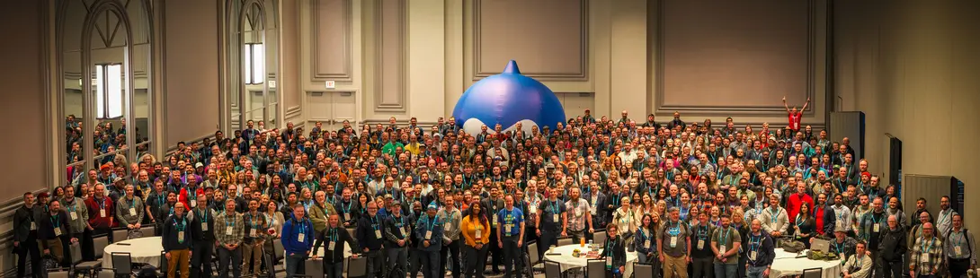 A wide-angle shot of a crowd of participants gathered in a Chicago conference hall for DrupalCon 2026, with a large, blue inflatable Druplicon mascot centered in the room.