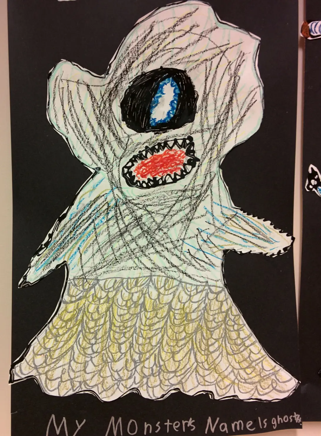 A child's drawing of a ghost-monster with one large blue eye and a wide red mouth full of teeth, on black paper. The caption reads 'My monster's name is ghost.'