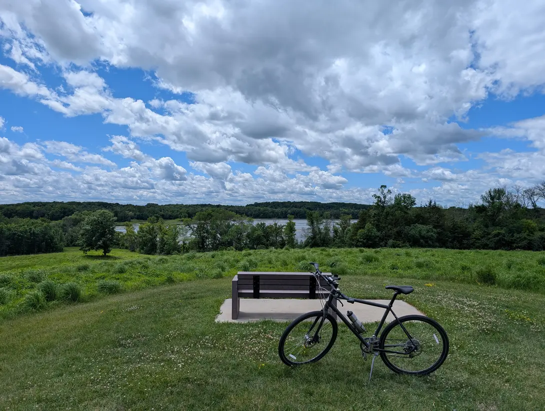 A dark grey bicycle on its kickstand in mown grass in front of an empty wooden bench on a concrete pad, with a meadow, a line of trees, a river, and tall clouds behind them.
