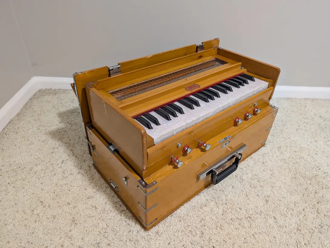 A wooden harmonium sitting open on beige carpet in the corner of a room, its top panel raised above the black and white keyboard, with a row of chrome and red stop knobs and a carrying handle along the front.