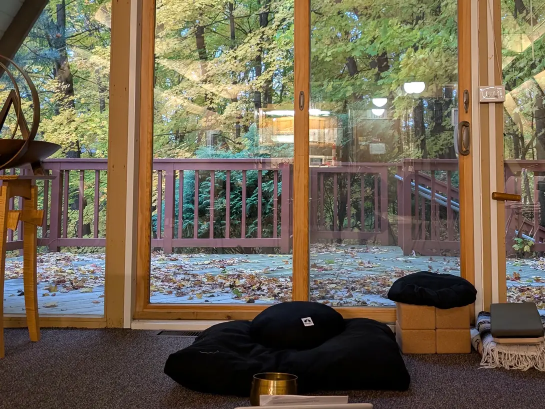 Meditation cushions, blocks, and a singing bowl on the floor of an A-frame room, in front of tall windows looking out at autumn trees and a leaf-covered deck.