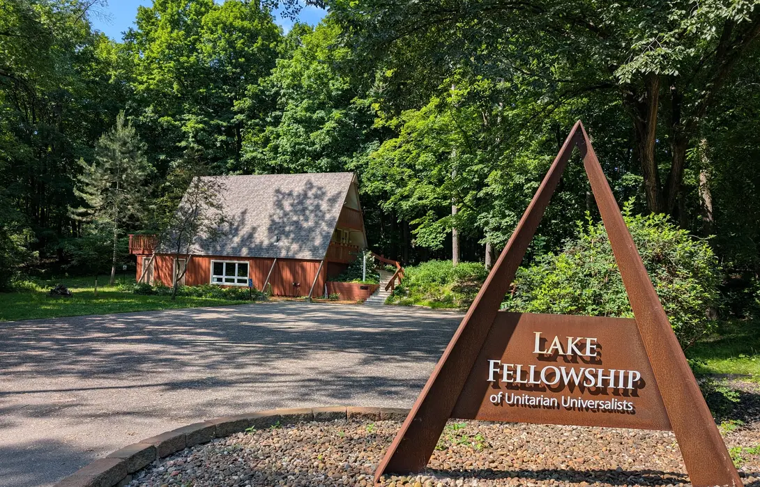 A triangular rusted-metal sign in the foreground reading "Lake Fellowship of Unitarian Universalists" with a wooden A-frame building nestled in lush green woods in the background.