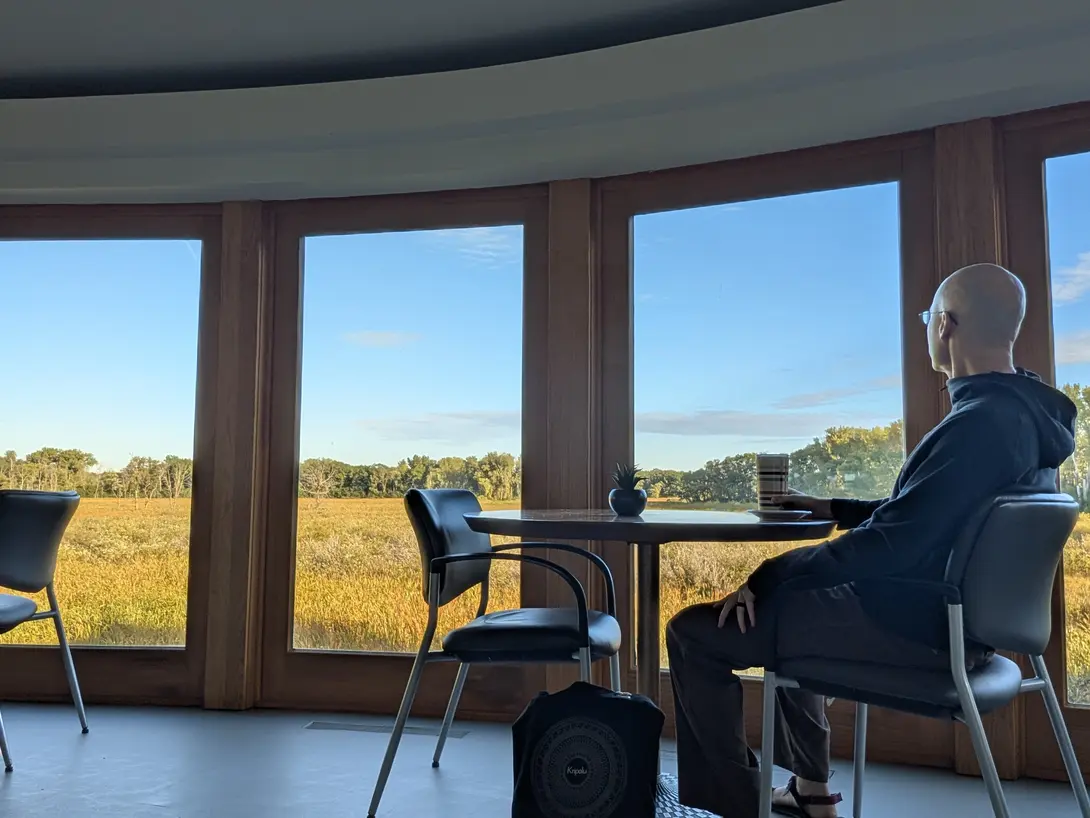 A person in a hoodie sits at a small round table by a curved bank of windows, mug in hand, looking out over a golden marsh toward a tree line under a clear sky.