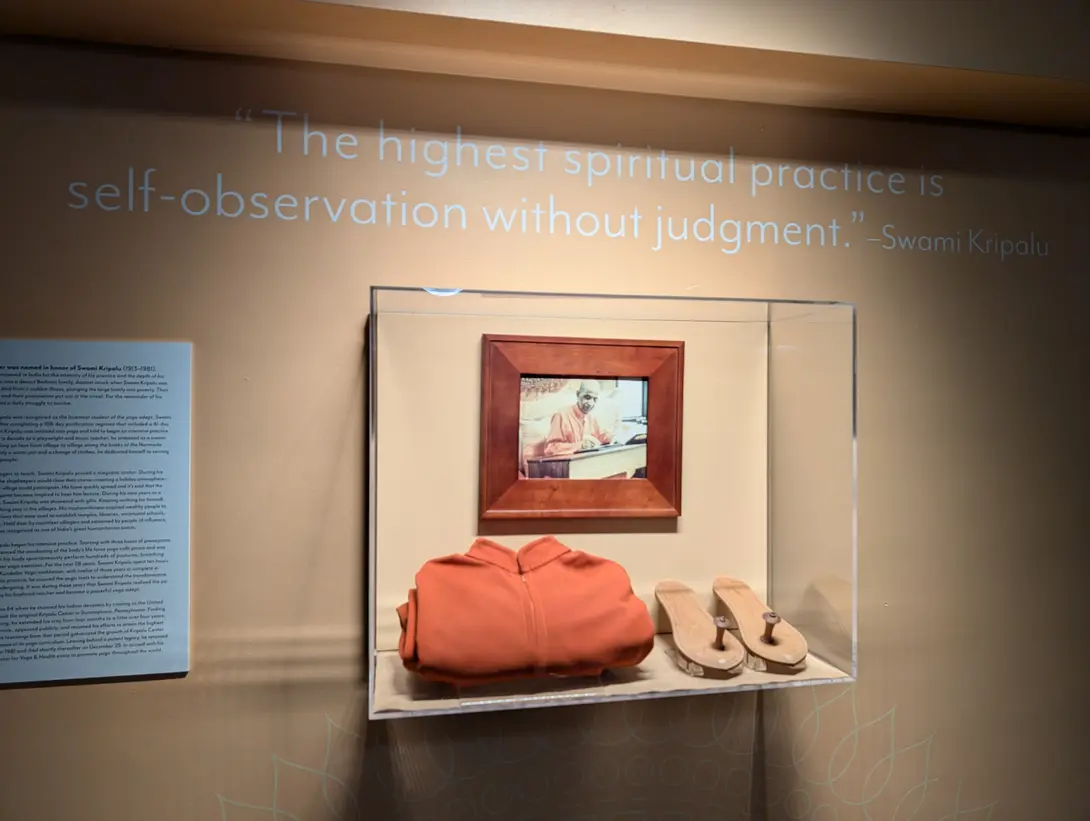 A quote painted on a wall reading "The highest spiritual practice is self-observation without judgment" above a glass display case holding a framed photo of Swami Kripalu at a writing desk, a folded orange robe, and a pair of wooden sandals, with a text panel to the left.