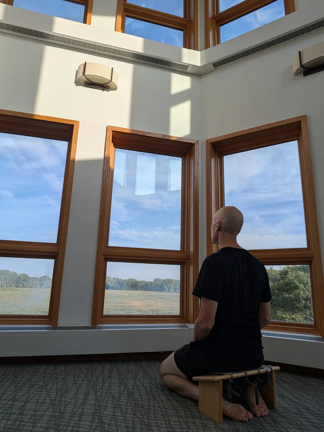 A bald man named Matthew in a black T-shirt and shorts kneels barefoot on a low wooden meditation bench, seen from behind, facing tall wood-framed windows that look out over a marsh and treeline under a blue sky, with sunlight falling across the white wall above.