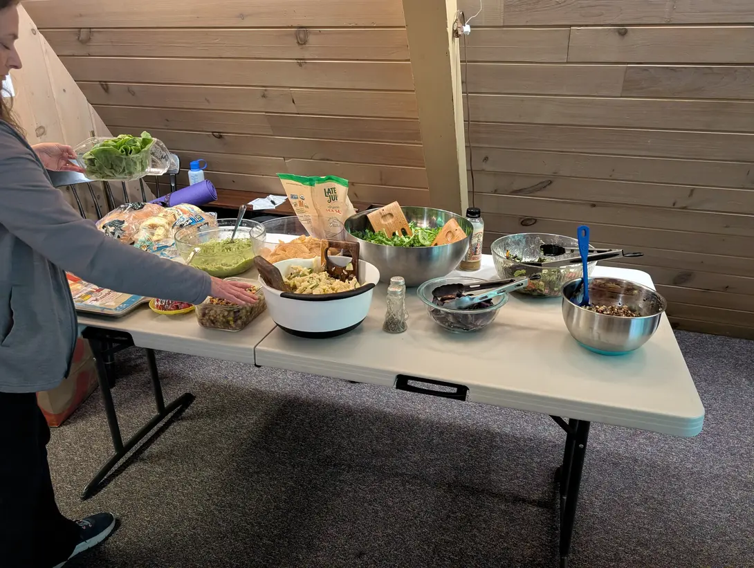 A woman sets a plastic container of lettuce onto a folding table crowded with potluck dishes, including guacamole, pasta salad, a large steel bowl of green salad, tortilla chips, bread, and several smaller bowls, with a rolled purple yoga mat on a chair behind her.