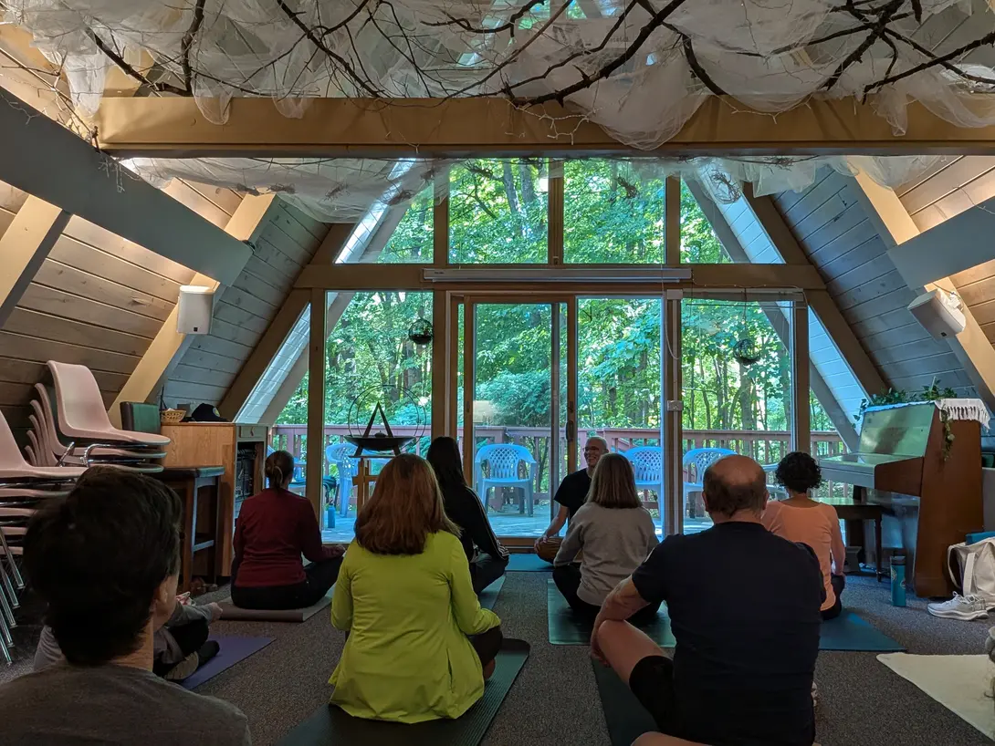 A group of people sit on yoga mats on the floor of an A-frame room, facing a bald man with glasses seated in front of tall windows, trees visible through the glass and gauzy fabric and bare branches hung across the ceiling.