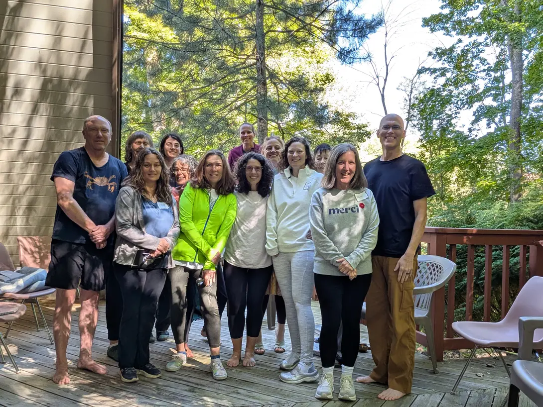 Thirteen people stand together on a wooden deck outside a small A-frame building, with trees behind them.