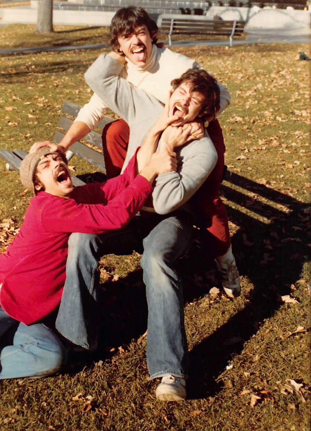 Three men beside a park bench, grabbing one another, sticking out their tongues, and making exaggerated faces.