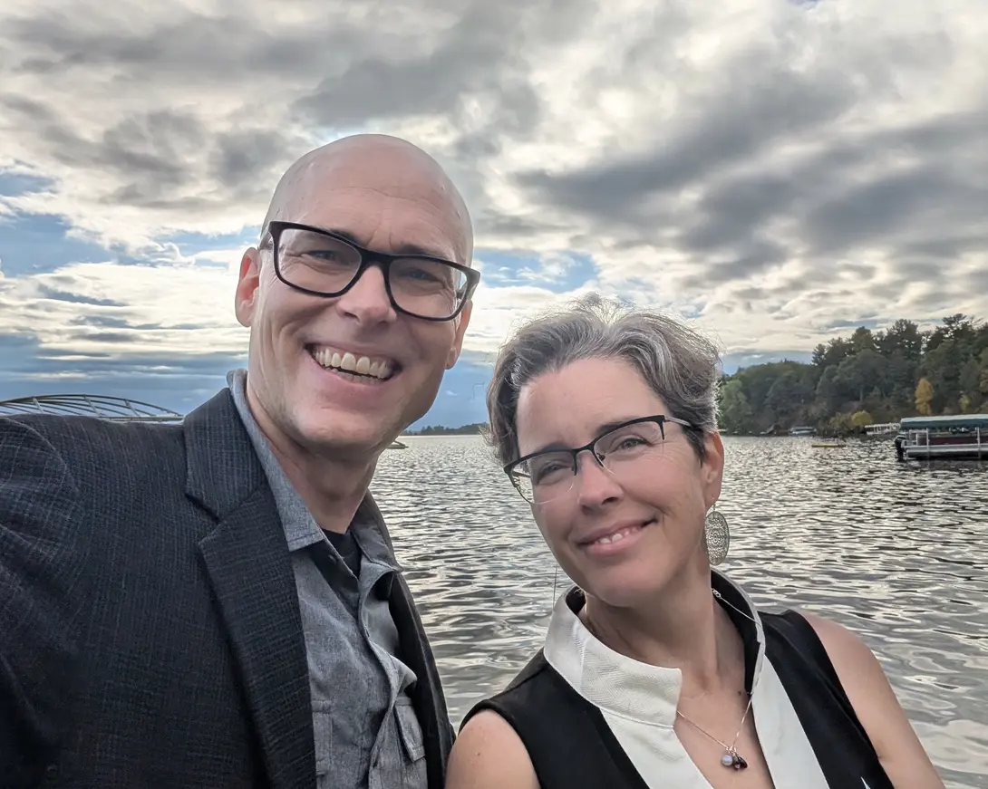 Matthew and Marnie smiling in a selfie on the shore of a lake, with a cloudy sky behind them and trees along the far bank